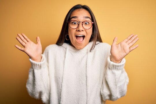 Young beautiful asian woman wearing casual sweater and glasses over yellow background celebrating crazy and amazed for success with arms raised and open eyes screaming excited. Winner concept