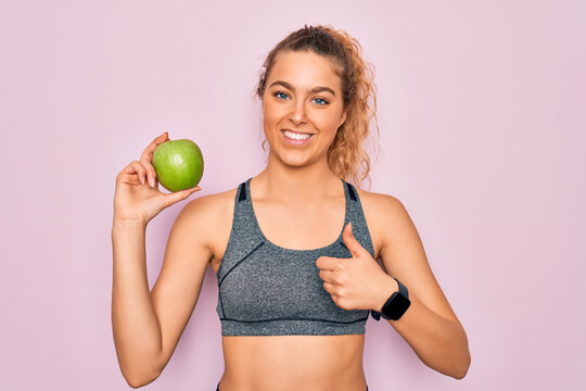 Young beautiful blonde sporty woman with blue eyes holding healthy green apple fruit happy with big smile doing ok sign, thumb up with fingers, excellent sign