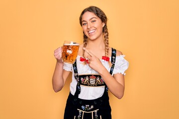 Beautiful blonde german woman with blue eyes wearing octoberfest dress drinking jar of beer very happy pointing with hand and finger