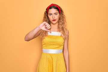 Beautiful blonde pin-up woman with blue eyes wearing diadem standing over yellow background looking unhappy and angry showing rejection and negative with thumbs down gesture. Bad expression.