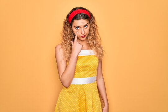 Beautiful blonde pin-up woman with blue eyes wearing diadem standing over yellow background Pointing to the eye watching you gesture, suspicious expression