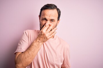 Fototapeta premium Middle age hoary man wearing casual t-shirt standing over isolated pink background smelling something stinky and disgusting, intolerable smell, holding breath with fingers on nose. Bad smell