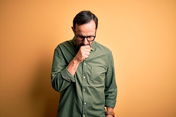 Middle age hoary man wearing casual green shirt and glasses over isolated yellow background feeling unwell and coughing as symptom for cold or bronchitis. Health care concept.