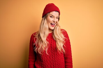 Young beautiful blonde woman wearing casual sweater and wool cap over white background winking...
