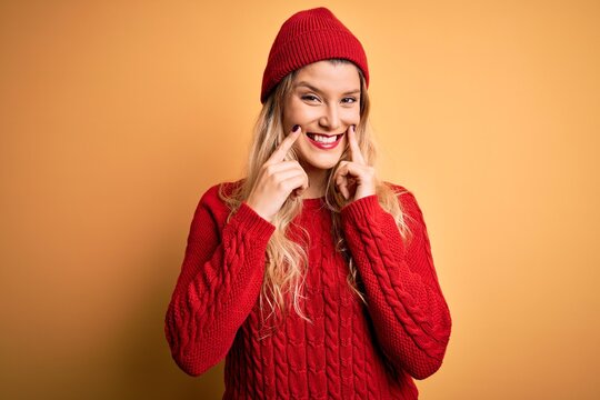 Young beautiful blonde woman wearing casual sweater and wool cap over white background Smiling with open mouth, fingers pointing and forcing cheerful smile