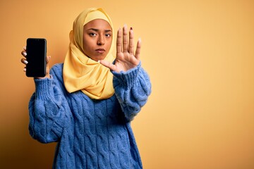 Young African American afro woman wearing muslim hijab showing smartphone with open hand doing stop sign with serious and confident expression, defense gesture