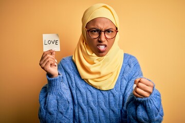 Young African American afro woman wearing muslim hijab holding love reminder paper message annoyed and frustrated shouting with anger, crazy and yelling with raised hand, anger concept