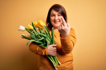 Beautiful plus size woman holding romantic bouquet of natural tulips flowers over yellow background Beckoning come here gesture with hand inviting welcoming happy and smiling