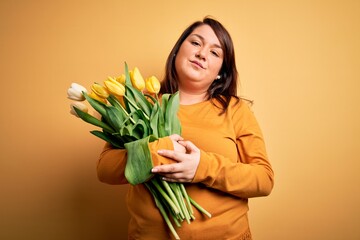 Beautiful plus size woman holding romantic bouquet of natural tulips flowers over yellow background looking sleepy and tired, exhausted for fatigue and hangover, lazy eyes in the morning.