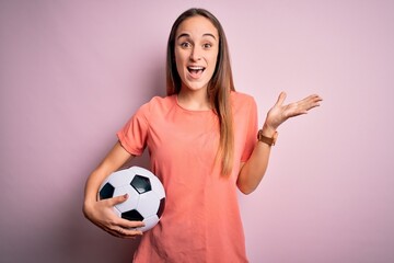 Young beautiful player woman playing soccer holding football ball  over pink background very happy and excited, winner expression celebrating victory screaming with big smile and raised hands