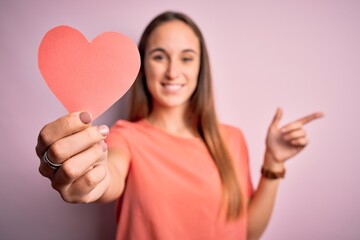 Young beautiful romantic woman holding paper heart shape over isolated pink background very happy pointing with hand and finger to the side