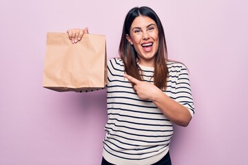 Young beautiful woman holding deliver paper bag with takeaway food over pink background smiling happy pointing with hand and finger