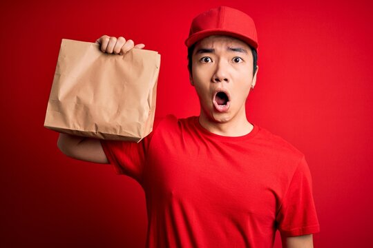 Young handsome chinese delivery man holding takeaway paper bag with food scared in shock with a surprise face, afraid and excited with fear expression