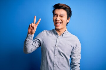 Young handsome chinese man wearing casual shirt standing over isolated blue background smiling with happy face winking at the camera doing victory sign with fingers. Number two.