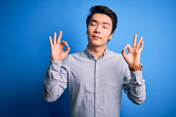 Fototapeta premium Young handsome chinese man wearing casual shirt standing over isolated blue background relaxed and smiling with eyes closed doing meditation gesture with fingers. Yoga concept.