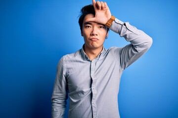 Young handsome chinese man wearing casual shirt standing over isolated blue background making fun of people with fingers on forehead doing loser gesture mocking and insulting.