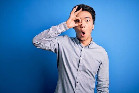 Young handsome chinese man wearing casual shirt standing over isolated blue background doing ok gesture shocked with surprised face, eye looking through fingers. Unbelieving expression.