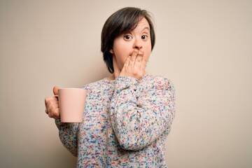 Young down syndrome woman drinking a cup of hot coffee over isolated background cover mouth with...
