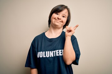 Young down syndrome volunteer woman wearing social care charity t-shirt cheerful with a smile on face pointing with hand and finger up to the side with happy and natural expression