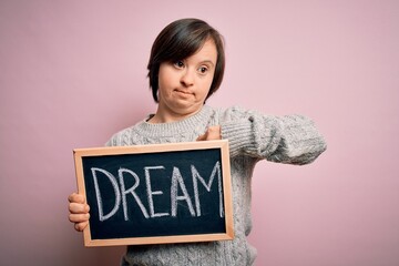 Young down syndrome woman holding blackboard with dream word as message of happiness with surprise face pointing finger to himself