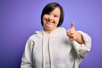 Young down syndrome sports woman wearing fitness sweatshirt over purple background doing happy thumbs up gesture with hand. Approving expression looking at the camera showing success.