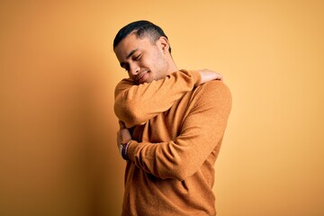 Young brazilian man wearing casual sweater standing over isolated yellow background Hugging oneself happy and positive, smiling confident. Self love and self care