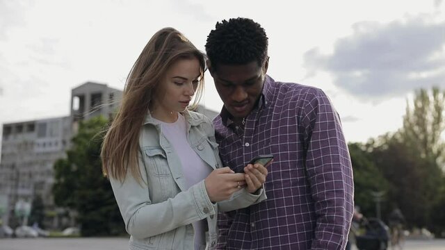 Two multiracial friends looking down into smartphones app or website in a city