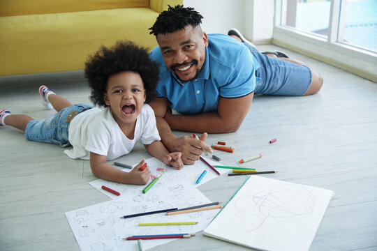 Black African family dad son together at home. Young father and little boy smile at camera and lying on warm floor and drawing coloring on book with color pencil crayon at living room