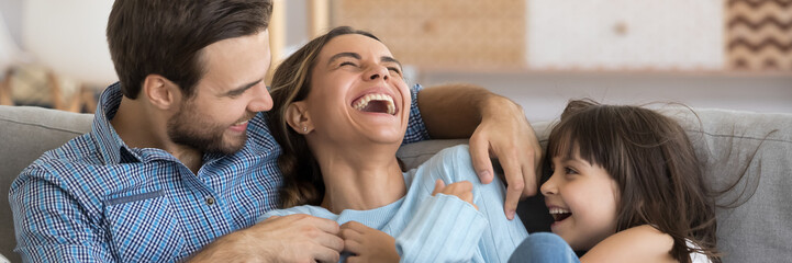 Laughing husband and daughter tickling mother family play together having fun resting on sofa at...
