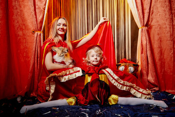 Family with dog during stylized theatrical circus photo shoot in beautiful red location. Model mother and daughter with small toy animal posing on stage with curtain