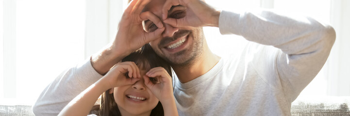 Fototapeta premium Smiling father and little daughter having fun at home, making fingers like glasses eyewear shape looking through binoculars smiling looking at camera. Horizontal photo banner for website header design