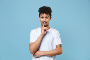 Fototapeta na wymiar Secret young african american guy in casual white t-shirt posing isolated on blue background studio. People lifestyle concept. Mock up copy space. Say hush be quiet with finger on lips shhh gesture.