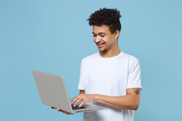 Smiling young african american guy in casual white t-shirt posing isolated on pastel blue wall background studio portrait. People lifestyle concept. Mock up copy space. Working on laptop pc computer.