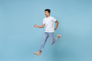 Fototapeta na wymiar Funny young african american guy in casual white t-shirt posing isolated on pastel blue wall background studio portrait. People emotions lifestyle concept. Mock up copy space. Jumping spreading hands.