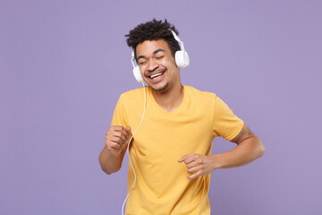 Cheerful young african american guy in casual yellow t-shirt isolated on violet wall background. People lifestyle concept. Mock up copy space. Listen music with headphones keeping eyes closed dancing.