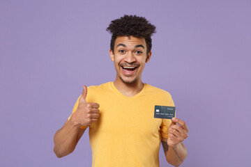 Excited young african american guy in casual yellow t-shirt posing isolated on pastel violet background studio. People lifestyle concept. Mock up copy space. Hold credit bank card, showing thumb up.