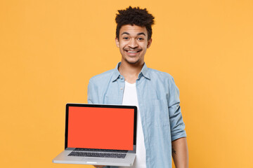 Smiling young african american guy in casual blue shirt posing isolated on yellow wall background studio. People lifestyle concept. Mock up copy space. Hold laptop pc computer with blank empty screen.