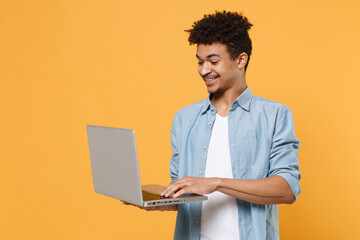 Smiling young african american guy in casual blue shirt posing isolated on yellow wall background studio portrait. People emotions lifestyle concept. Mock up copy space. Working on laptop pc computer.
