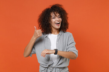 Funny young african american woman in gray casual clothes isolated on orange background studio portrait. People lifestyle concept. Mock up copy space. Wearing smart watch on hand, showing thumb up.