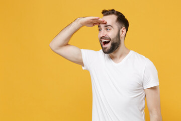 Excited young bearded man guy in white casual t-shirt posing isolated on yellow background in studio. People lifestyle concept. Mock up copy space. Holding hand at forehead looking far away distance. - obrazy, fototapety, plakaty