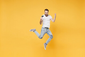 Joyful young bearded man guy in white casual t-shirt posing isolated on yellow wall background studio. People lifestyle concept. Mock up copy space. Jumping hold credit bank card doing winner gesture.