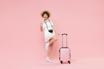 Joyful tourist girl in summer dress hat with photo camera suitcase isolated on pink background. Female traveling abroad to travel weekend getaway. Air flight journey concept. Doing winner gesture.