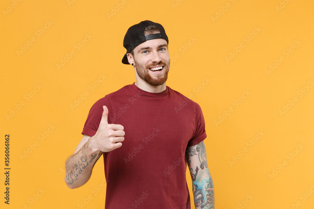 Wall mural smiling young bearded tattooed man guy in casual t-shirt, black cap posing isolated on yellow wall b