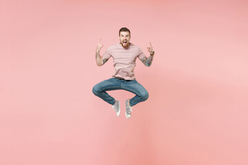 Crazy young tattooed man guy in pastel casual t-shirt posing isolated on pink background studio. People lifestyle concept. Mock up copy space. Jumping depicting heavy metal rock sign horns up gesture.