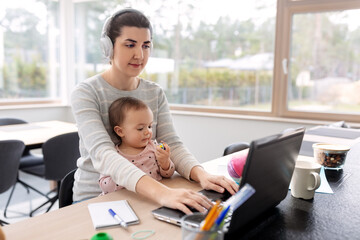 remote job, multi-tasking and family concept - happy in headphones mother with baby working on laptop at home office