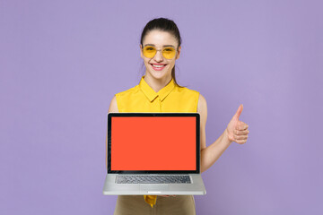 Smiling young brunette woman girl in yellow casual shirt isolated on violet background. People lifestyle concept. Mock up copy space. Hold laptop pc computer with blank empty screen, showing thumb up.