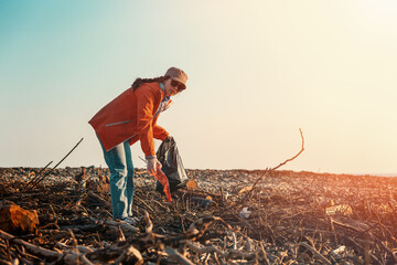 A young Caucasian female volunteer in a jacket collects garbage on a dirty beach. In the...