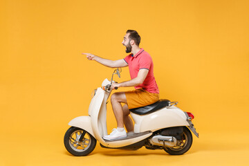Fototapeta premium Side view of shocked young bearded man guy in casual summer clothes driving moped isolated on yellow background. Driving motorbike transportation concept. Mock up copy space. Point index finger aside.