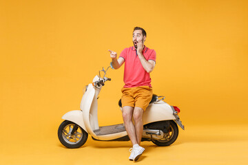 Fototapeta na wymiar Shocked young bearded man guy in casual summer clothes standing near moped isolated on yellow wall background. Driving motorbike transportation concept. Pointing index finger aside, put hand on cheek.