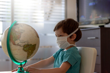 Image of cute little toddler with white medical mask,sitting at home in quarantine and looking at globe.Self-isolation and social distance.Close up of a boy looking at the globe with a protective mask